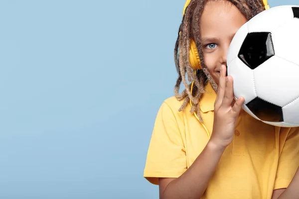 Smiling little boy with african dreads cover face with soccer ball over blue background