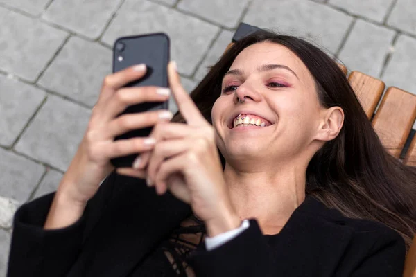 一个笑着的年轻女子躺在长椅上看智能手机的画像 — 图库照片