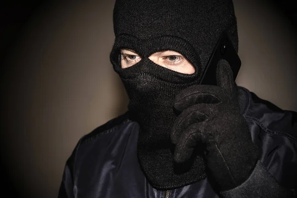 An extortionist in black clothes with a balaclava covering his face. A cruel fraudster uses a mobile phone. Mobile racket. Telephone fraud with bank accounts. — Stock Photo, Image