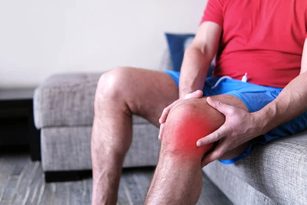 Artritida je onemocnění kloubů. Muž na gauči, mačkající si koleno před nesnesitelnou bolestí. — Stock fotografie