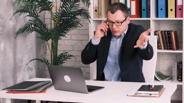 Недовольный разочарованный бизнесмен в очках и пиджаке, разговаривающий по мобильному телефону, сидящий в офисе на полках с папками и злой жестикулирующей глядя в монитор открытого ноутбука — стоковое видео