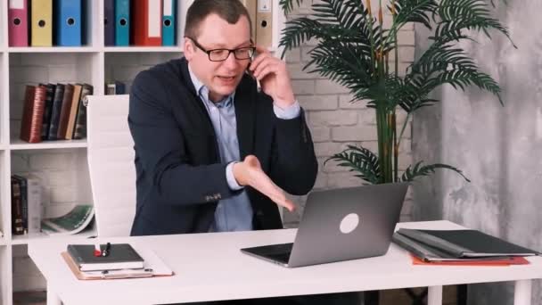 Empresario frustrado descontento en gafas y chaqueta hablando en el teléfono móvil sentado en la oficina en el escritorio mirando en el monitor de un ordenador portátil abierto. Movimiento lento — Vídeos de Stock