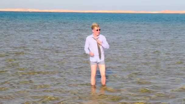 Um homem engraçado com uma camisa, shorts, meias e óculos de sol ajusta sua gravata, de pé no mar, segurando um telefone celular em suas mãos em um dia ensolarado de verão. Downshifter, freelancer. — Vídeo de Stock