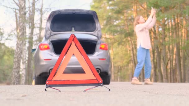 Figyelmeztető vörös háromszög. Szürke autó villogó vészvilágítással az út mentén egy baleset után. A csomagtartó nyitva van. A nő próbál mobil kapcsolatot találni, hogy felhívja a vontatót. Lassú mozgás, 4k — Stock videók