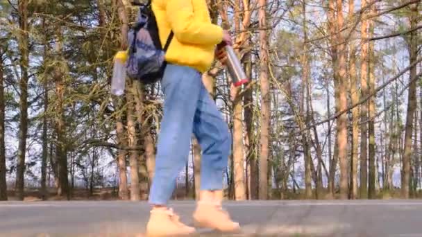 Автостопом концепция путешествия. Молодая туристка с рюкзаком в желтой куртке с термосом в руках ходит по дороге вдоль сосновой рощи. Медленное движение, 4k — стоковое видео
