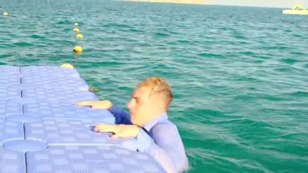 Um jovem alegre salta da água e senta-se no cais com uma camisa e cuecas, jogando uma gravata molhada sobre o ombro. Movimento lento, 4k — Vídeo de Stock