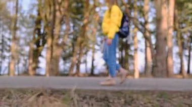Otostop konsepti. Sarı ceketli, elinde termosla sırt çantalı genç bir turist çam koruluğu boyunca yol boyunca yürüyor. Seçici odaklanma. Yavaş çekim, 4k