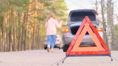 Kırmızı üçgen uyarısı. Elinde telefonu olan üzgün genç bir kadın yoldan geçtikten sonra koşuyor. Yol boyunca açık bagajlı, yanıp sönen acil durum ışığı olan gri bir araba. Yardım bekliyorum. Ağır çekim, 4K. Araba arızası