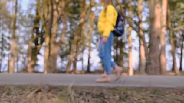 Hitchwalking reisconcept. Jonge toerist met een rugzak in een geel jasje met een thermosfles in haar handen loopt langs de weg langs een dennenbos. Selectieve focus. Langzame beweging, 4k — Stockvideo