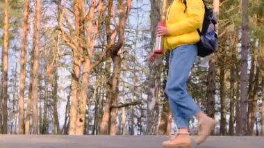 Otostop konsepti. Sarı ceketli, elinde termosla sırt çantalı genç bir turist çam koruluğu boyunca yol boyunca yürüyor. Seçici odaklanma. Yavaş çekim, 4k