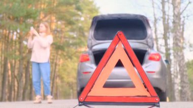 Kırmızı üçgen uyarısı. Kazadan sonra yol boyunca yanıp sönen acil durum ışığı olan gri bir araba. Bagaj açık. Bir kadın arabanın yanında yürür ve çekici çağırmak için mobil bir bağlantı bulmaya çalışır..