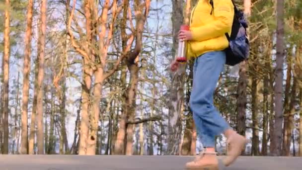 ヒッチハイキング旅行のコンセプト。彼女の手の中に魔法瓶と黄色のジャケットのバックパックを持つ若い女性の観光客は、松林に沿って道路に沿って歩いています。選択的フォーカス。スローモーション4k — ストック動画
