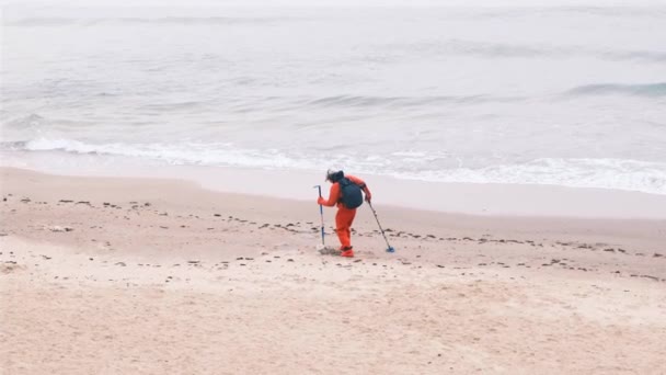 Člověk chodí s detektorem kovu v písku. Elektronický detektor kovu a kopeček. Muž hledá luxus, který turisté ztratili ve vodě a na pláži. Pobřeží, zpěněné vlny. 4k, zpomalený pohyb — Stock video