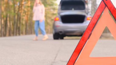 Araba arızası. Genç bir kadın telefonda konuşurken bozuk bir arabanın yanında yol boyunca yürüyor. Acil durum üçgenine yakın plan, odağını kaybetmiş bir kadın. Araba alarmı yanıp sönüyor. Yavaş çekim, 4k