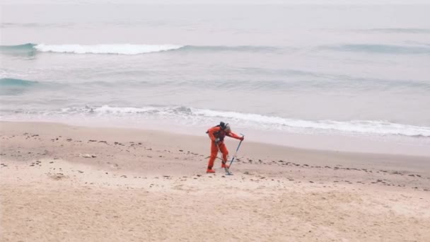 Kırmızı tulumlu bir adam bulutlu bir sonbahar gününde plajda elektronik metal dedektörlü kayıp lüksleri arıyor. Kumu bir kepçeyle kazıyor ve hazineyi aramak için eliyor. 4k — Stok video