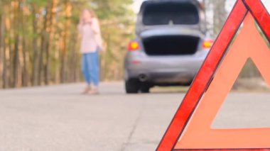 Araba arızası. Genç bir kadın telefonda konuşurken bozuk bir arabanın yanında yol boyunca yürüyor. Acil durum üçgenine yakın plan, odağını kaybetmiş bir kadın. Araba alarmı yanıp sönüyor. Yavaş çekim, 4k