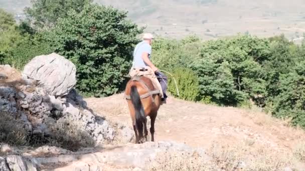 Pria senior menunggang kuda di jalan pegunungan. Pemandangan dari belakang seorang pria tua dengan topi menuruni jalan batu. 4k — Stok Video