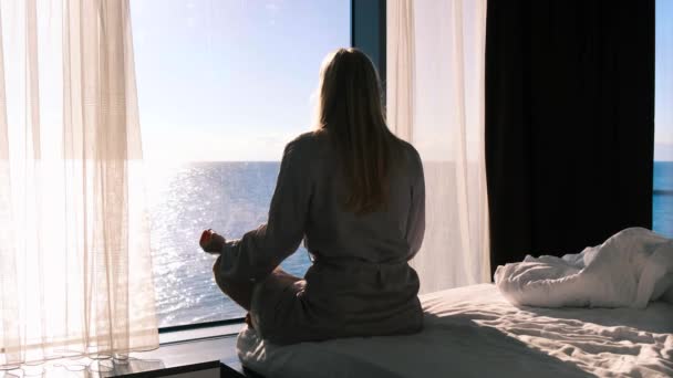 Medytacja i zdrowie psychiczne. Widok z tyłu medytującej blondynki siedzącej na krawędzi łóżka naprzeciwko panaramskiego okna z widokiem na morze w słoneczny dzień. 4k, w czasie rzeczywistym — Wideo stockowe