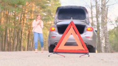 Kırmızı üçgen uyarısı. Kazadan sonra yol boyunca yanıp sönen acil durum ışığı olan gri bir araba. Bagaj açık. Bir kadın arabanın yanında yürür ve çekici çağırmak için mobil bir bağlantı bulmaya çalışır..