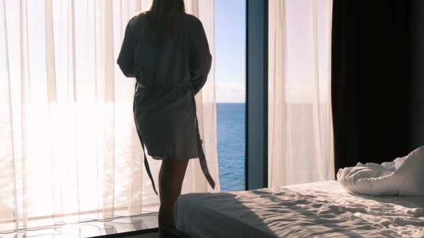 灰色の絹のバスローブのブロンドは、窓、海と太陽の景色にカーテンを開きます。スリムな女性は、パノラマの窓の外を見て、ローブのベルトを結び、ベッドの上に横たわっています。スローモーション。4k — ストック動画