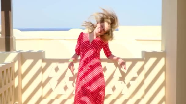 Young beautiful happy woman in a red dress in white polka dots smiles and plays with her hair looking into the camera, standing on the terrace. Vacation and summer holidays concept. — Stock Video