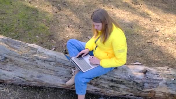 Werk of studie op afstand. Zicht op de vrouw met een laptop zittend in het bos op een omgevallen boom in een geel jasje en spijkerbroek. 4k, slow motion — Stockvideo