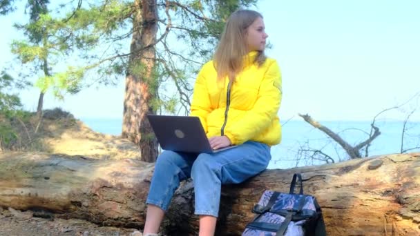 Aantrekkelijke student gebruikt een laptop zittend op een boomstam in het bos. Het meisje drukt, draait haar hoofd, ademt de frisse lucht diep genietend van de natuur en keert terug naar haar werk of studie. 4k, slow motion — Stockvideo