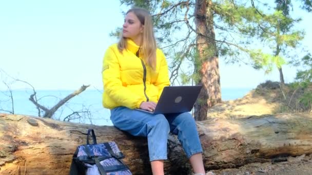Aantrekkelijke student gebruikt een laptop zittend op een boomstam in het bos. Het meisje drukt, draait haar hoofd, ademt de frisse lucht diep genietend van de natuur en keert terug naar haar werk of studie. 4k — Stockvideo