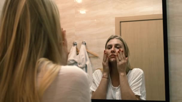 Уставшая женщина наносит питательный или увлажняющий крем на лицо, глядя в стоячее зеркало в ванной комнате. Уход за собой, косметические процедуры. 4k, slow motion — стоковое видео