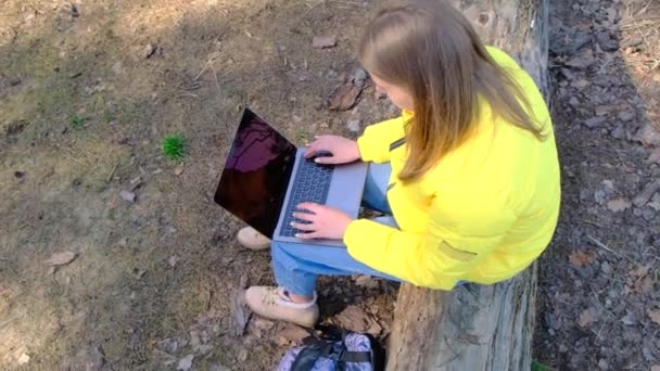Werk of studie op afstand. Bovenaanzicht van de vrouw met behulp van een laptop zittend op een boomstam in een geel jasje en spijkerbroek. Meisje typt snel. Langzame beweging, 4k — Stockvideo