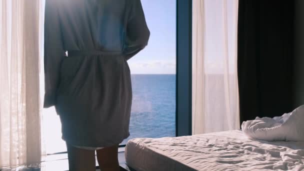 Klidná šťastná mladá blondýnka v županu si vychutnává svou ranní kávu, která sedí na posteli na pozadí panoramatického okna s výhledem na moře za slunečného dne. Zpomal, 4k. Selektivní zaměření — Stock video