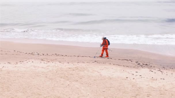 Muž s detektorem kovu kráčí po pláži v červeném obleku. Zpomalený pohyb, 4k. — Stock video