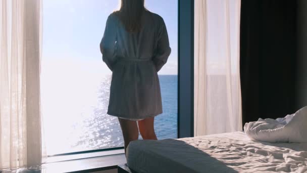 バスローブを着た穏やかな幸せな若いブロンドは、晴れた日にパノラマの窓を背景に海が立っている美しい景色を楽しんでいます。スローモーション、 4k 。選択的焦点. — ストック動画