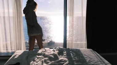 Bornozlu mutlu genç sarışın güneşli bir günde panoramik pencerenin önünde duran deniz manzarasının tadını çıkarıyor. Kadın ellerini kaldırıyor. Ağır çekim, 4K. Seçici odak.