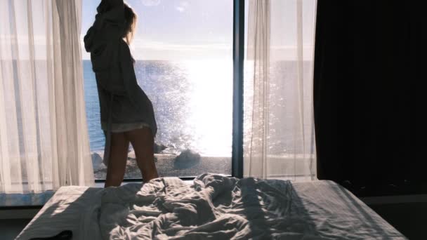 バスローブを着た穏やかな幸せな若いブロンドは、晴れた日にパノラマの窓を背景に海の美しい景色を楽しんでいます。女性は手を伸ばしていた。スローモーション、 4k 。選択的焦点. — ストック動画