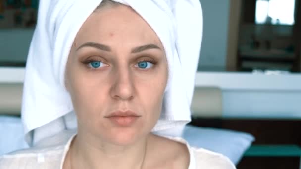 Portrait of a young woman with a towel on her head after a shower. Blue-eyed girl looks into the camera adjusting the towel on her head. She sits on the bed in a white robe. — Stockvideo