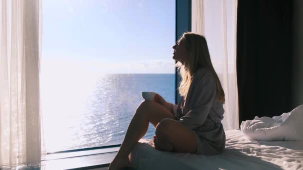 Mujer rubia feliz pacífica en albornoz disfruta de su café de la mañana sentado en su cama contra el telón de fondo de la ventana panorámica con vistas al mar en un día soleado. A cámara lenta, 4k. Enfoque selectivo — Vídeo de stock