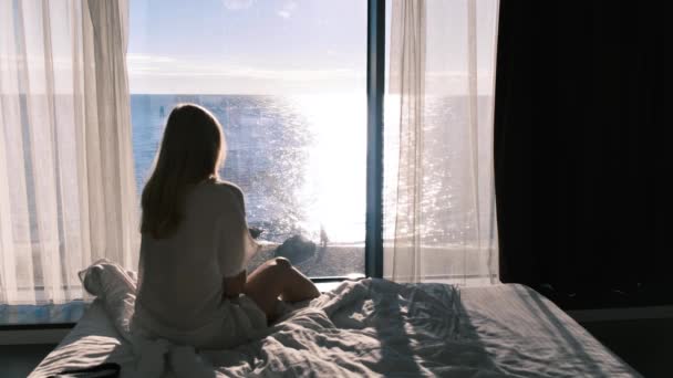 晴れた日に海を見下ろすパナルム窓の反対側のベッドに座っている女性の後部ビュー。女性はベッドから出て、フレームを残します。スローモーション4k — ストック動画