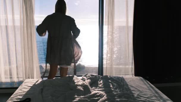 晴れた日には海を見下ろすパノラマの窓から見えるローブを着た女性がいます。寝室の窓の前に立つ目覚めたての少女の後ろ姿。スローモーション4k — ストック動画
