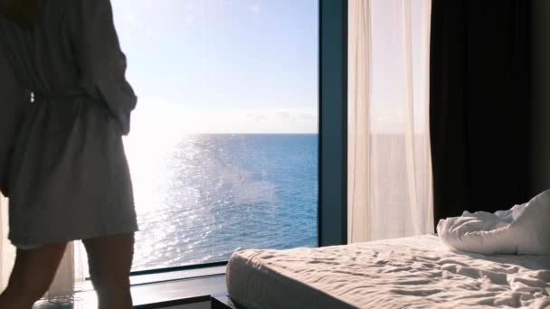 Egy csendes, boldog fiatal szőke, fürdőköpenyben élvezi a reggeli kávé áll a háttérben egy panorámás ablakban, kilátással a tengerre egy napsütéses napon. Lassabban, 4k. Szelektív fókusz. — Stock videók