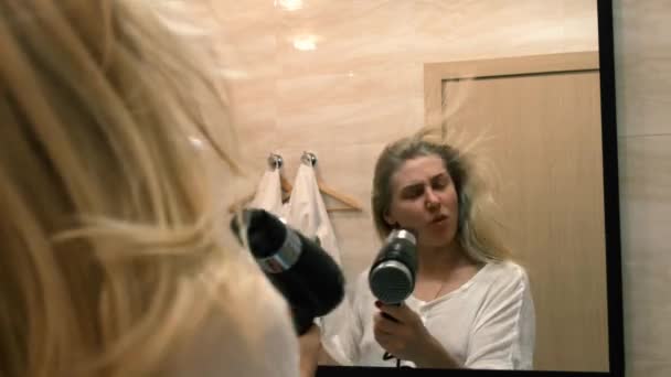 Jonge vrouw danst en zingt in een werkende haardroger voor een spiegel in de badkamer. Haar fladdert in de wind. 4k, slow motion — Stockvideo