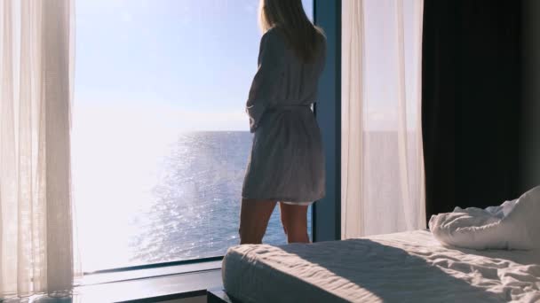 バスローブを着た穏やかな幸せな若いブロンドは、晴れた日に海を見下ろすパノラマの窓を背景に彼女の朝のコーヒースタンドを楽しんでいます。スローモーション、 4k 。選択的焦点. — ストック動画