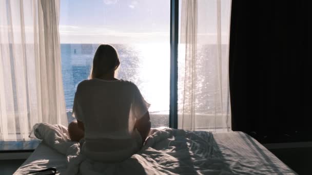 Hátul egy nő ül egy ágyon szemben egy panarum ablak kilátással a tengerre egy napsütéses napon. A nő felkel az ágyból, és elhagyja a keretet. Lassú mozgás, 4k — Stock videók