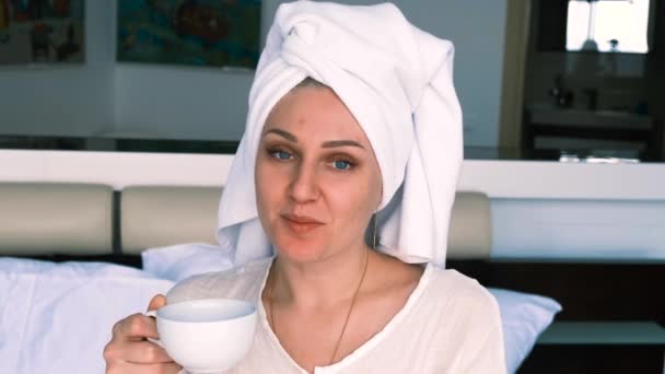 Glada flicka i badrock och handduk dricker kaffe medan du sitter på sängen och tittar på kameran. 4k, slow motion — Stockvideo
