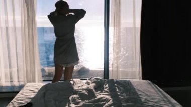 Bornozlu mutlu genç bir sarışın güneşli bir günde panoramik bir pencerenin önünde duran güzel bir deniz manzarasının keyfini çıkarıyor. Ağır çekim, 4K. Seçici odak.