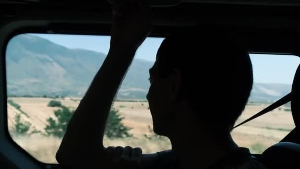 Viaje por carretera. Silueta de un hombre montado en un coche en el asiento del pasajero, disfrutando de una hermosa vista de las montañas. El hombre lleva puesto un cinturón de seguridad. — Vídeos de Stock