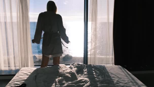 Спокойная счастливая молодая блондинка в халате наслаждается прекрасным видом на море, стоящее на фоне панорамного окна в солнечный день. Медленное движение, 4К. Селективный фокус. — стоковое видео