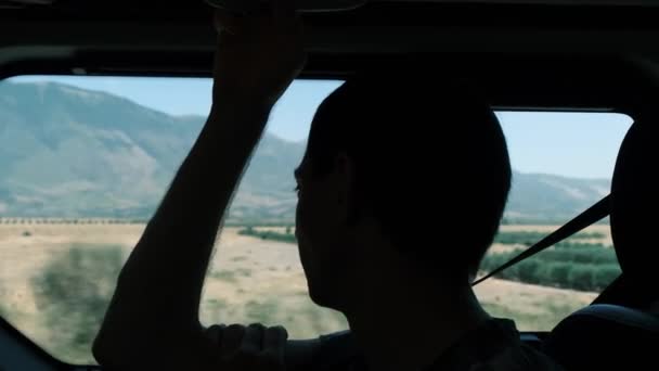 Viaje por carretera. Silueta de un hombre montado en un coche en el asiento del pasajero, disfrutando de una hermosa vista de las montañas. El hombre lleva puesto un cinturón de seguridad. — Vídeos de Stock