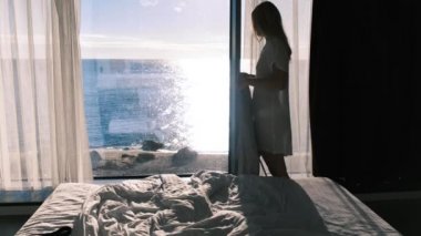 Bornozlu mutlu genç bir sarışın güneşli bir günde panoramik bir pencerenin önünde duran güzel bir deniz manzarasının keyfini çıkarıyor. Ağır çekim, 4K. Seçici odak.