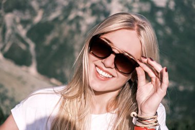 Kendine güvenen, gülümseyen, gülümseyen, güneş gözlüğü takan genç bir kadın, güneşli bir günde doğanın arka planına bakıyor. Mutlu kız gülümsüyor.